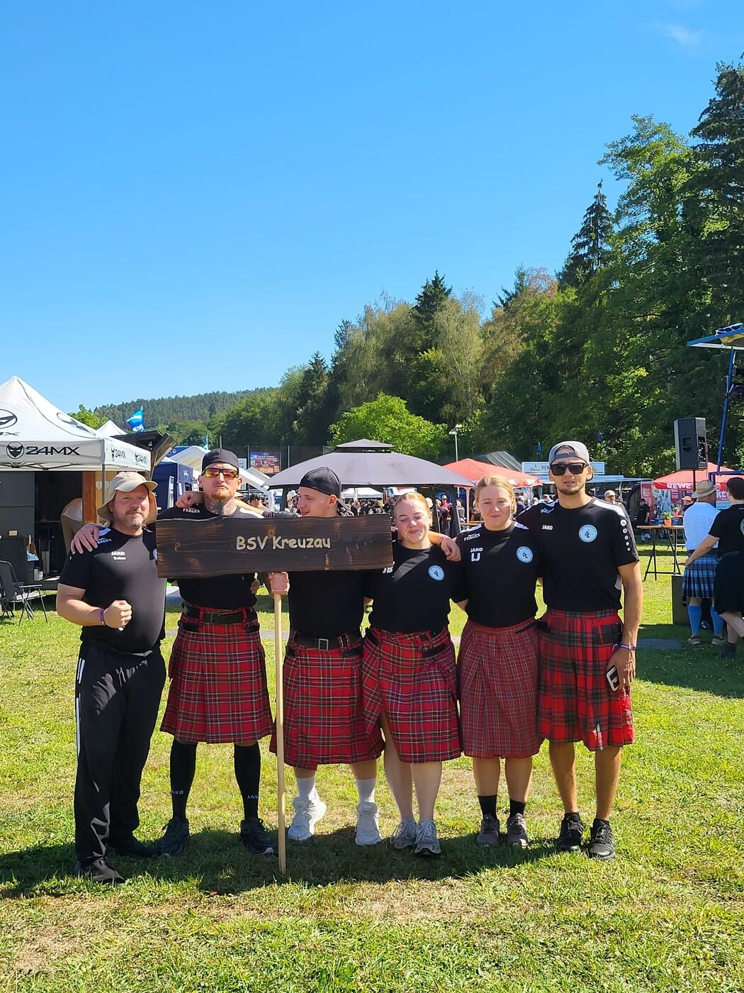 Das Team des BSV Kreuzau 2023 e.V. bei den 10. Highland Games am Stausee Obermaubach: sechs Sportlerinnen und Sportler in Vereinsshirts und Kilts hinter dem hölzernen Vereinsschild
